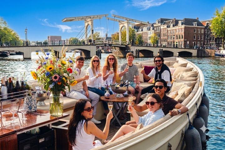 Group of people on a boat in a canal, toasting with drinks, bridge in background.