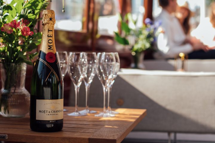 Champagne bottle and glasses on a table with blurred people in a bright room.