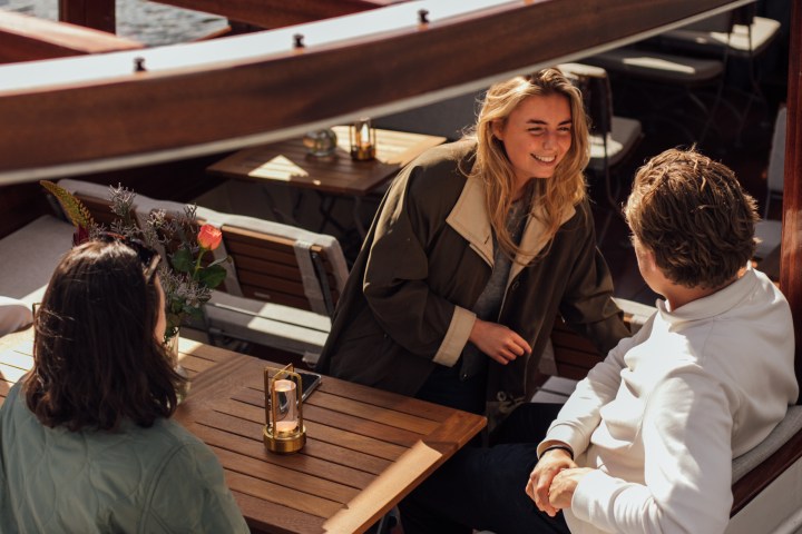 Three people chatting on a wooden boat with a table and flowers.