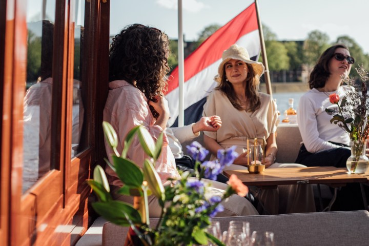 Three people sitting on a boat with drinks, flowers, and a flag in the background.
