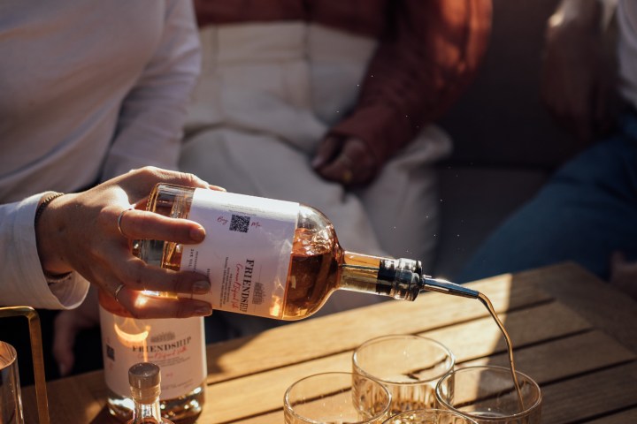 Person pouring whiskey into glasses on a wooden table by the water.