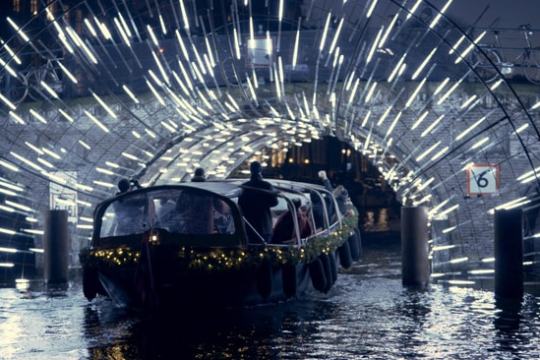 Boat passing under a canal bridge with illuminated lights forming an arch at night.