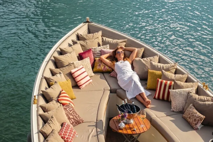 Woman in white dress relaxing on boat with cushions, table, drinks, and water view.