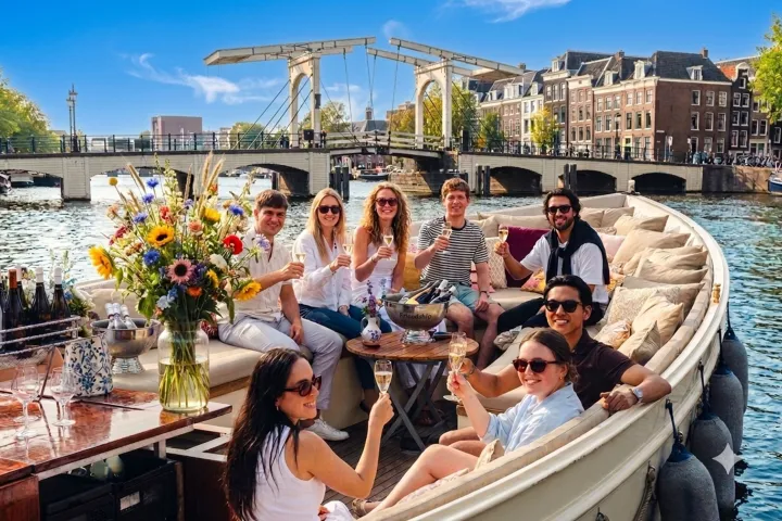 People on a boat toasting with drinks, canal and bridge in background.