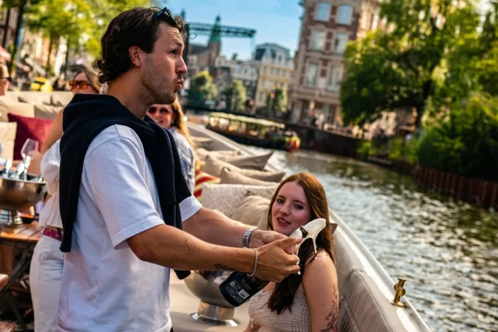 People on a boat by a canal in a city, one man holding a bottle.