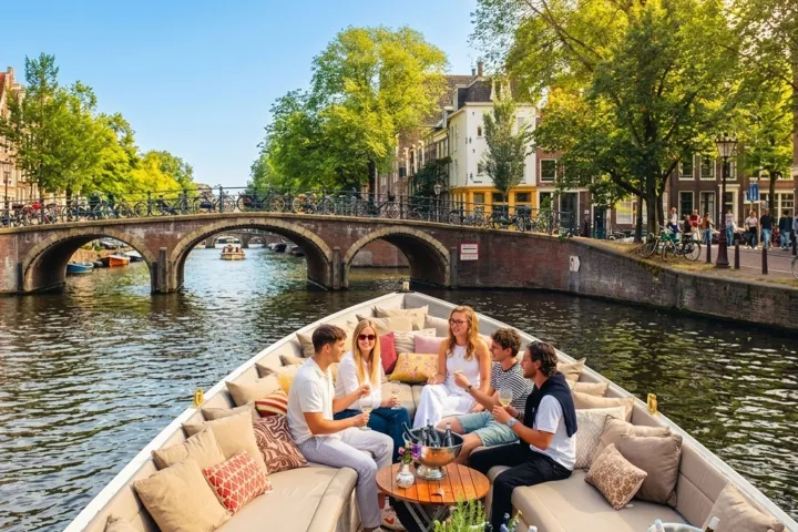 Group relaxing on a boat in a scenic canal with arched bridge and trees.